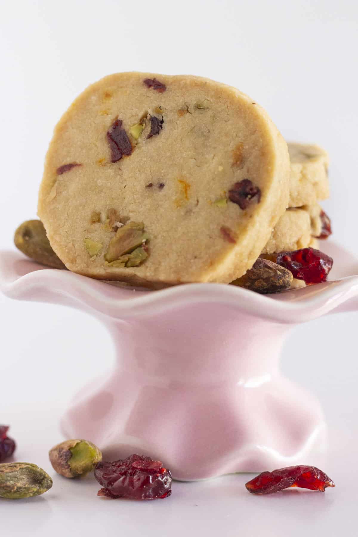 Biscuits shortbread aux canneberges et pistaches avec du zeste d'orange sur une petite assiette rose.