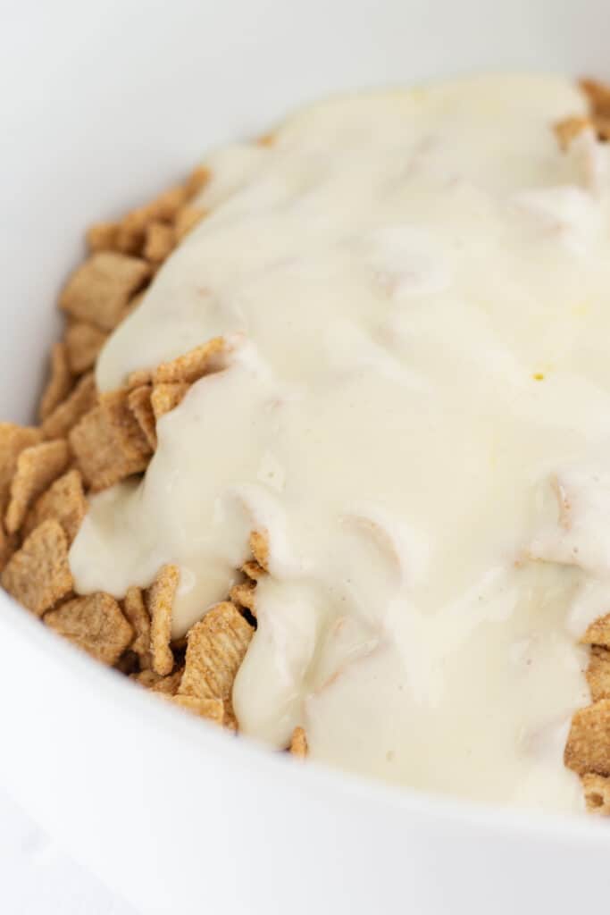 Melted marshmallows and butter poured over cinnamon toast crunch cereal in a white mixing bowl.