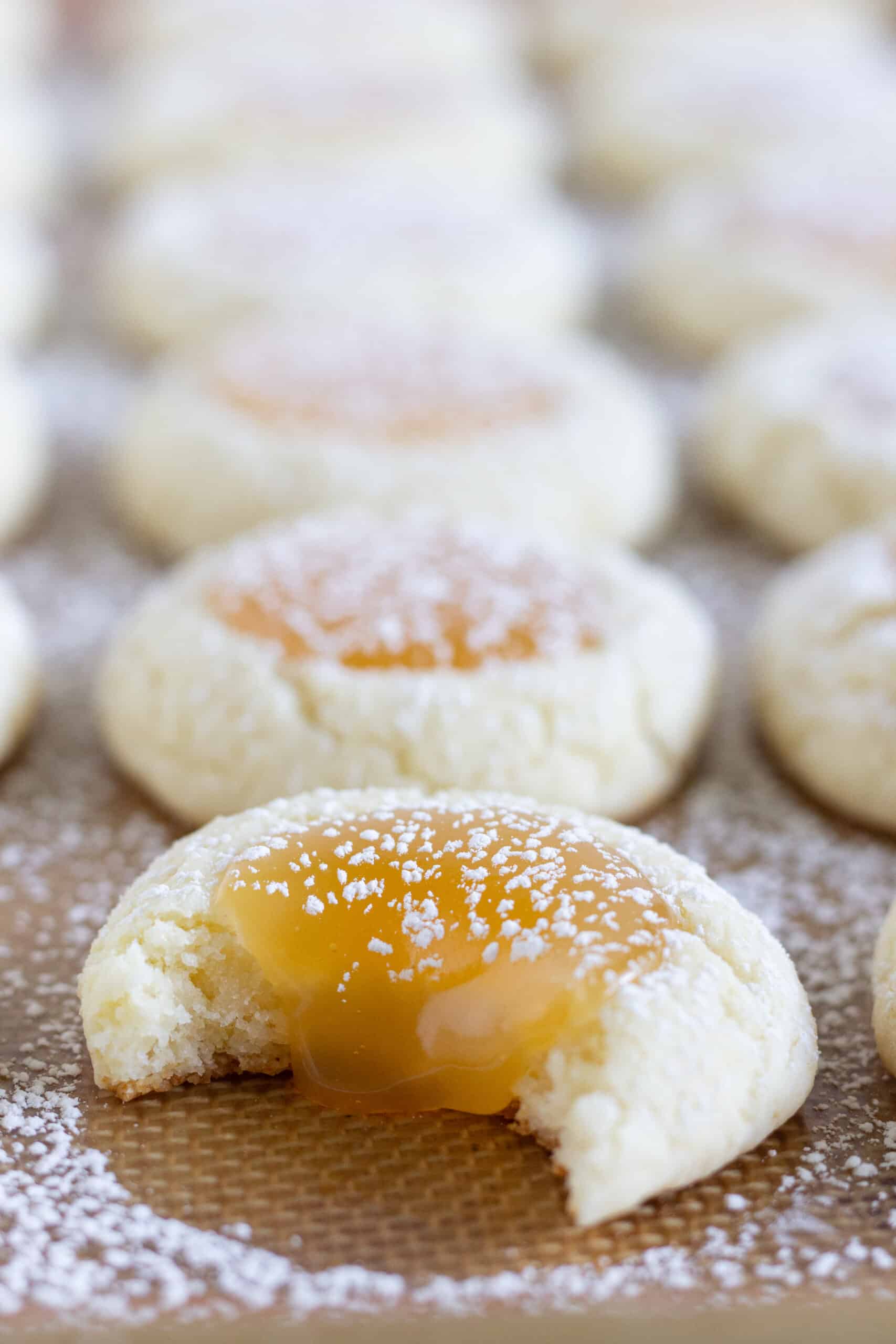 Lemon Curd Thumbprint Cookies on a baking sheet that are sprinkled with powdered sugar and one of the cookies have a bite taken out of it.