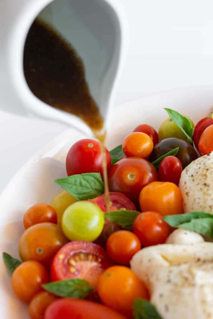 Balsamic vinaigrette pouring out of a pitcher onto a burrata caprese salad.