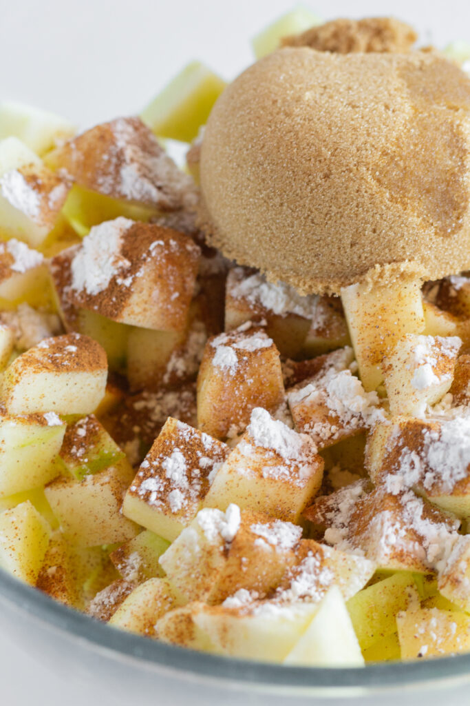 Apples, flour, brown sugar, and cinnamon in a glass mixing bowl.