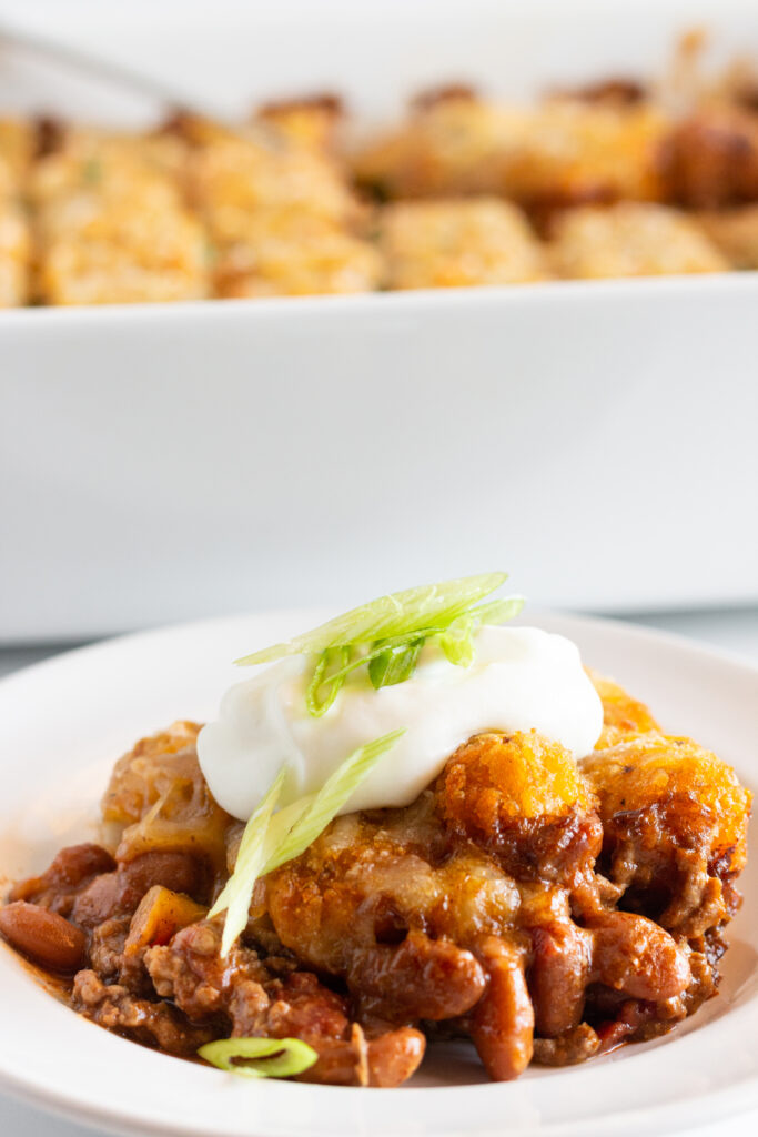 A serving of Chili Cheese Tater Tot Casserole with sour cream and green onion toppings and the casserole in the background.