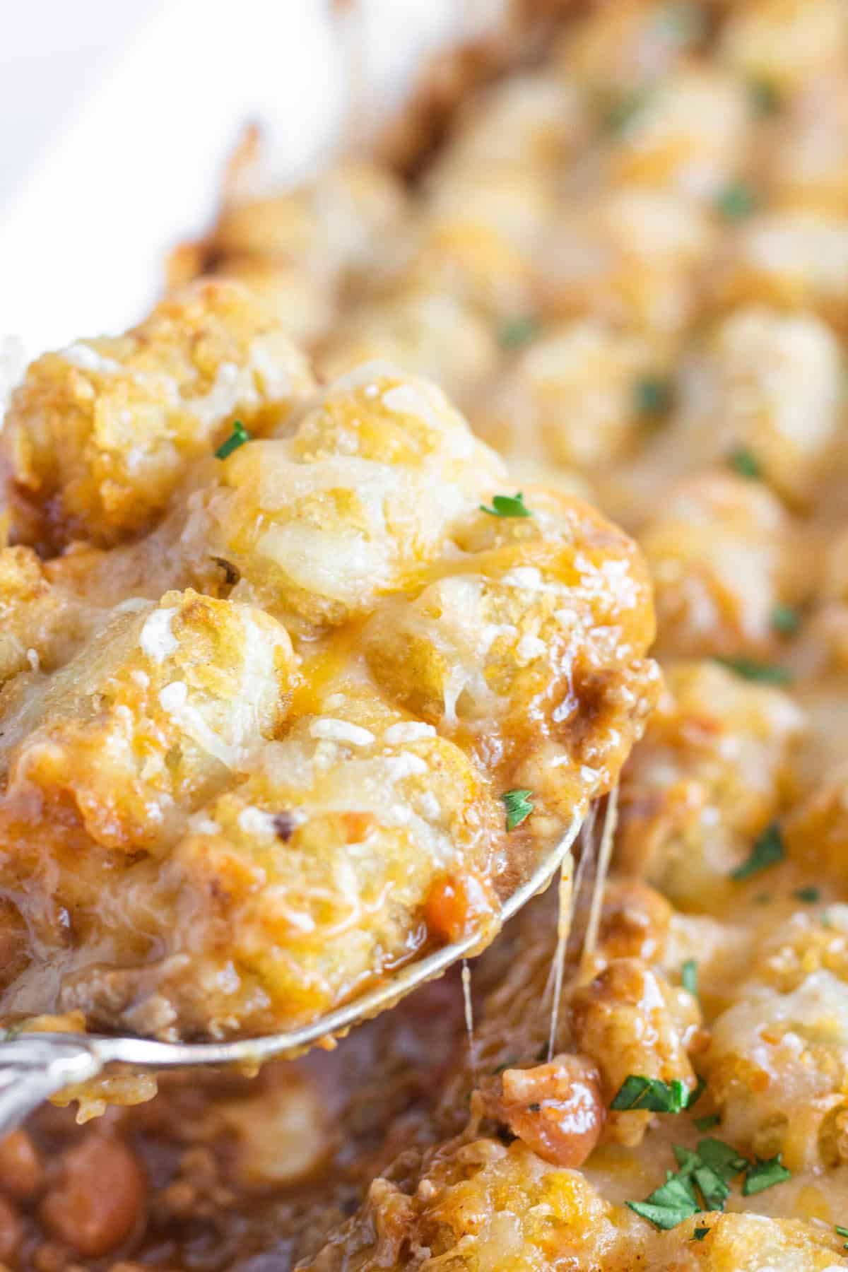 A spoon serving a helping of Chili Tater Tot Casserole with cheese pulling from the pan.