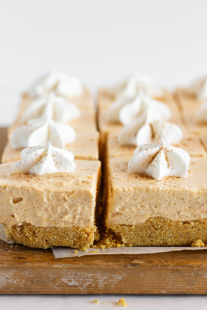 Pumpkin cheesecake bars on a wooden cutting board, topped with whipped cream.