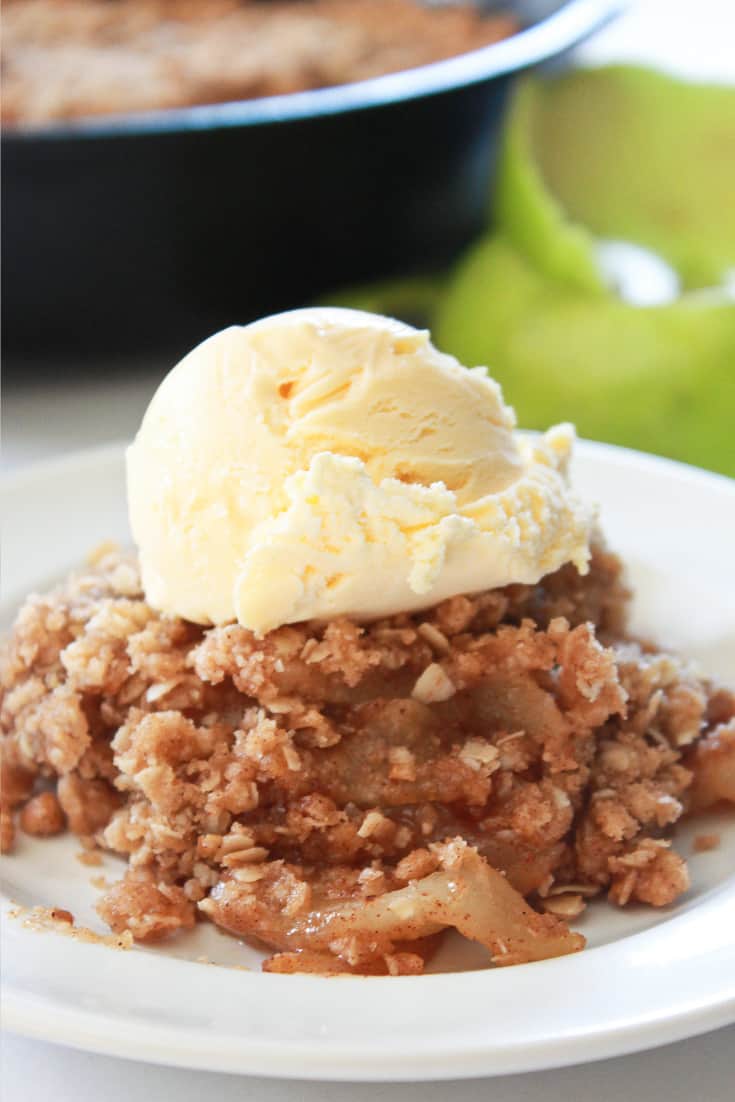 A serving of Skillet Apple Crisp with a scoop of ice cream and the skillet with the crisp in the back.