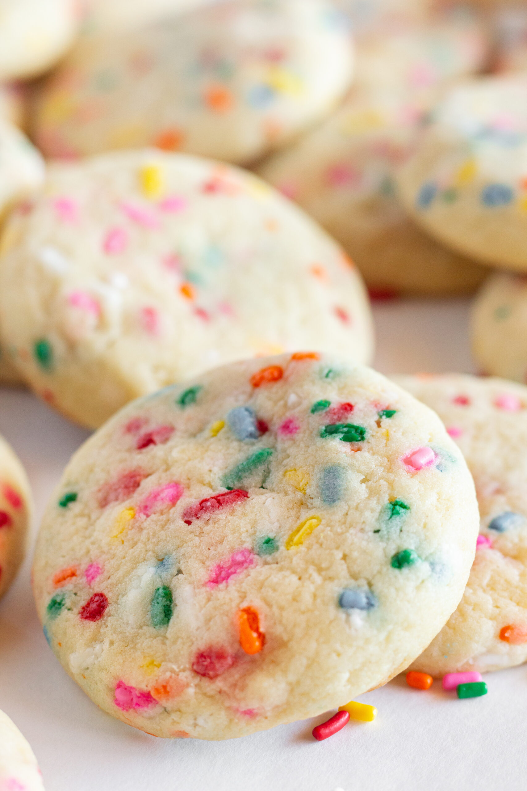 Sprinkle Sugar Cookies are stacked on a large white tray with extra colored sprinkles around them.