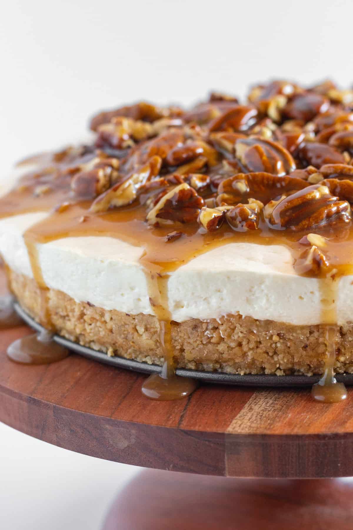 A whole Pecan Pie No Bake Cheesecake on a wood serving plate with the pecan topping dripping onto the plate.