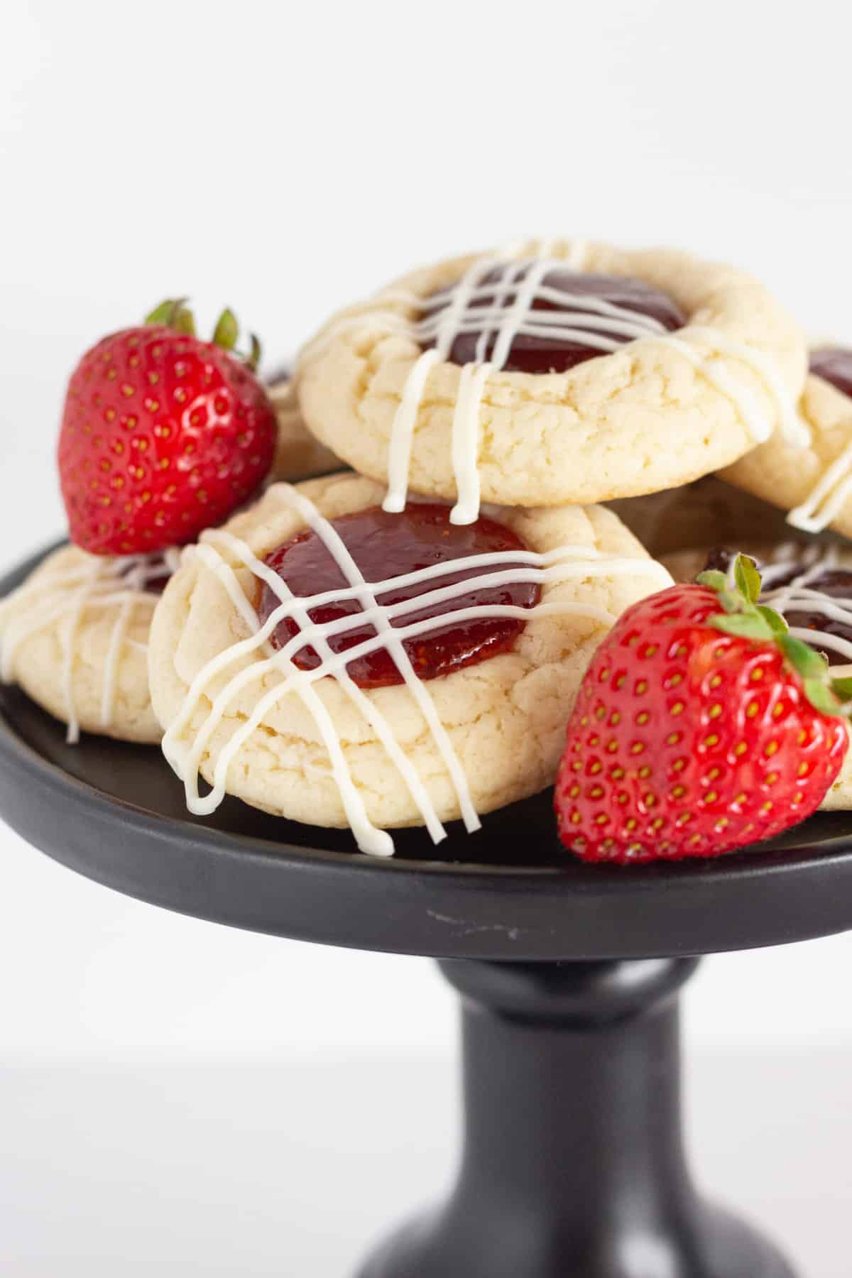 Easy Strawberry Jam Cookies are on a black cake plate.  The cookies are drizzled with while chocolate and a couple of fresh strawberries are on the plate.