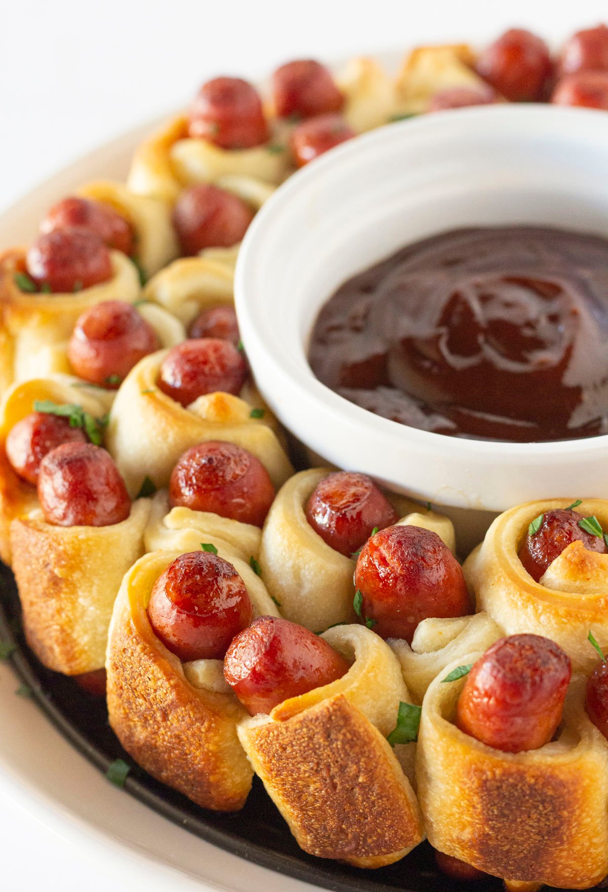Pigs in a blanket baked into a wreath shape with a bowl of bbq sauce in the middle.