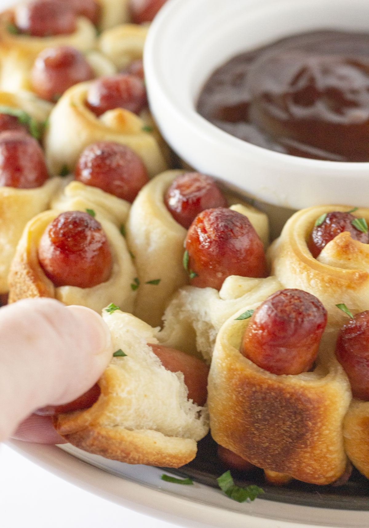 Pigs in a Blanket Wreath with bbq sauce in the bowl for dip and someone pulling one of the pigs and blanket from the wreath.