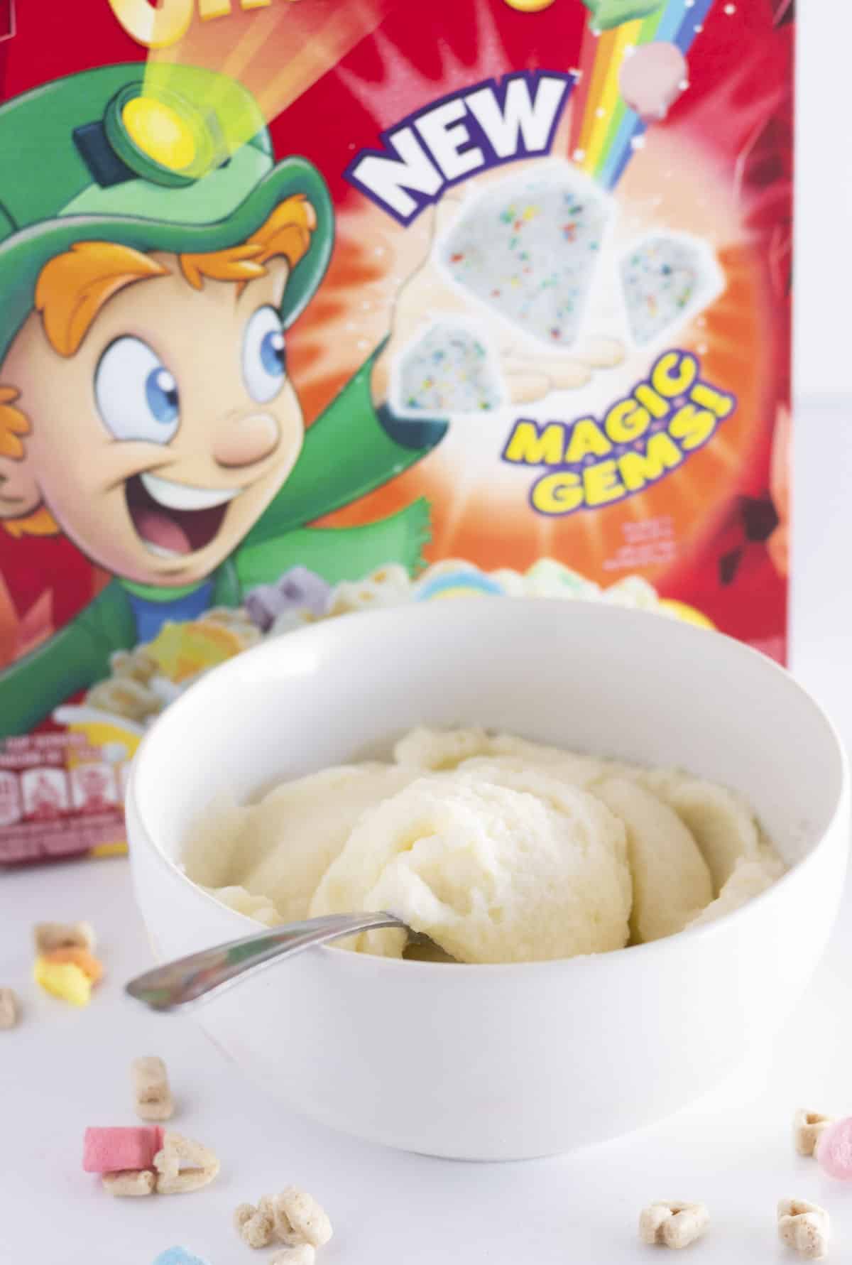 A bowl of cereal milk frosting with Lucky Charms in the background.
