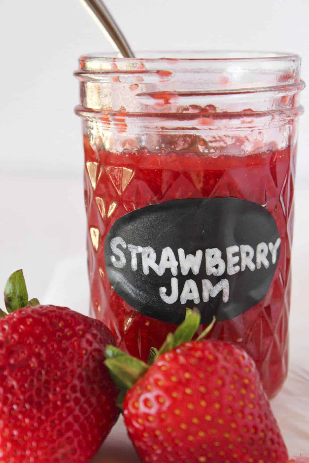 A jar of homemade strawberry jam with a spoon in the jar and fresh strawberries.